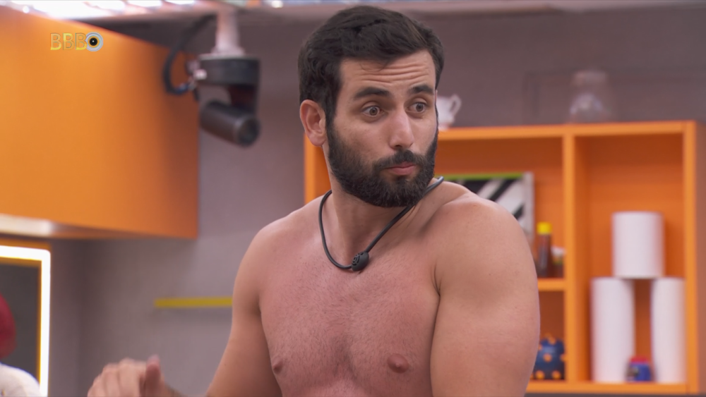 A imagem mostra Matteus Amaral, ex-participante do Big Brother Brasil (BBB). Ele está sem camisa, exibindo um físico musculoso e usa uma corda ou colar preto em volta do pescoço. Matteus tem cabelo escuro e barba cheia. No fundo, há uma estante laranja com diversos objetos e um armário de cor similar. O ambiente parece ser uma cozinha ou área comum da casa do BBB, com uma câmera de vigilância visível no canto superior esquerdo da imagem.