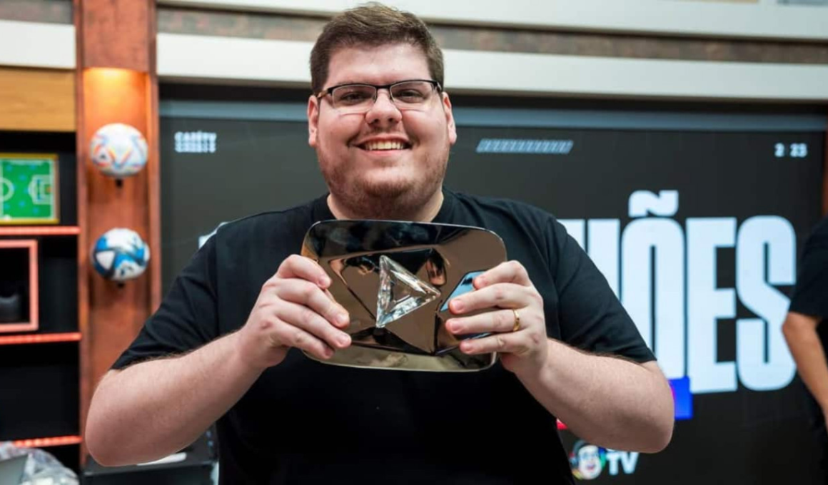 Casimiro Miguel, criador da CazéTV, sorrindo e segurando placa do YouTube em estúdio esportivo durante gravação de conteúdo digital.