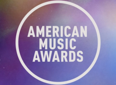 Logo do American Music Awards em destaque sobre fundo com tons degradê em roxo, azul e rosa. O nome da premiação aparece em letras brancas maiúsculas dentro de um círculo branco centralizado.