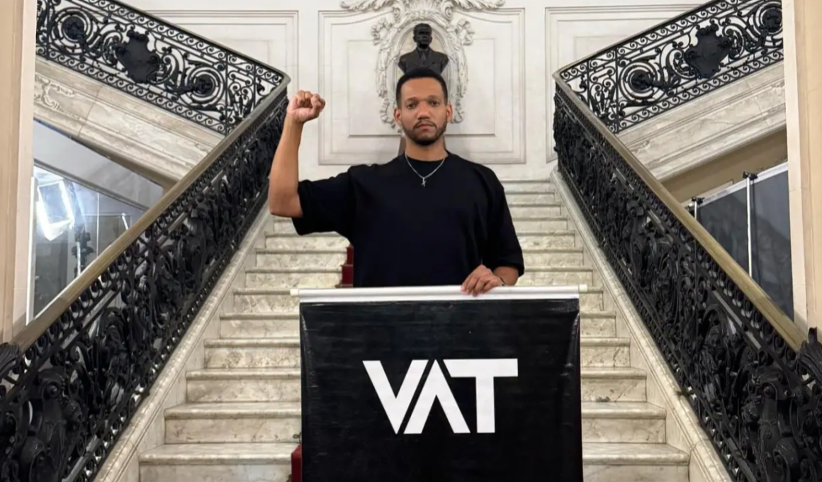 Rick Azevedo, um homem vestido com camiseta preta levanta o punho em escadaria de prédio histórico durante manifestação sobre o fim da escala 6x1, com banner escrito “VAT” em destaque.