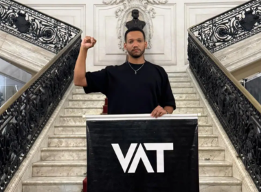 Rick Azevedo, um homem vestido com camiseta preta levanta o punho em escadaria de prédio histórico durante manifestação sobre o fim da escala 6x1, com banner escrito “VAT” em destaque.