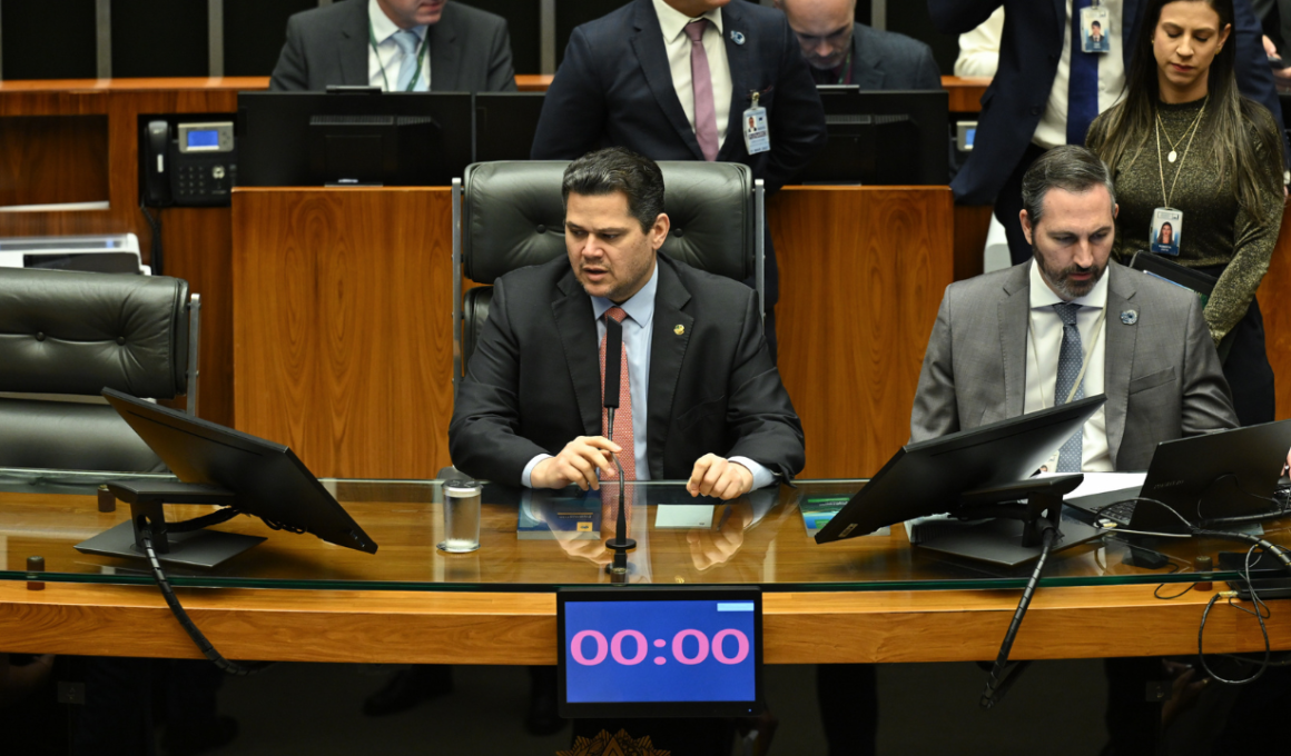 Imagem mostra Davi Alcolumbre presidindo sessão no Senado Federal em Brasília. O parlamentar aparece na mesa diretora do Congresso Nacional cercado por assessores e outros políticos durante debate envolvendo a CPI do Banco Master. A cena retrata o ambiente político institucional brasileiro, com destaque para o plenário e os equipamentos utilizados nas sessões legislativas.