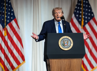 O presidente dos Estados Unidos, Donald Trump, aparece discursando em um púlpito oficial com o selo presidencial. Ele veste terno azul-marinho e gravata azul clara, enquanto duas bandeiras dos Estados Unidos estão posicionadas ao fundo. Trump gesticula com as mãos abertas durante o pronunciamento em um ambiente interno com cortinas brancas.