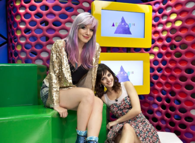 Imagem promocional do programa Acesso MTV mostra as apresentadoras MariMoon e Titi Müller em um estúdio vibrante e colorido da MTV Brasil. MariMoon aparece sentada em um bloco verde, usando jaqueta dourada com brilho e cabelo em tons de roxo e lilás, característica marcante de seu estilo alternativo. Ao lado, Titi Müller posa sorridente, com vestido estampado e visual jovem, apoiada de forma descontraída. O cenário ao fundo é composto por painéis geométricos em cores intensas como rosa, amarelo e azul, com telas exibindo o logotipo do programa, reforçando a identidade visual pop e moderna da MTV. A composição transmite leveza, estilo e conexão com o público jovem, características centrais do programa musical exibido entre 2009 e 2013.