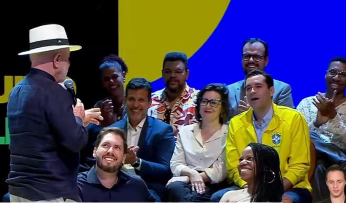 Presidente Lula, de costas e usando chapéu Panamá, discursa no lançamento do programa Tela Brasil. No palco, uma plateia sorridente acompanha o evento, incluindo um homem vestindo a jaqueta amarela da Seleção Brasileira de Futebol. Ao fundo, um painel azul e amarelo.