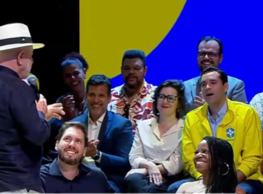 Presidente Lula, de costas e usando chapéu Panamá, discursa no lançamento do programa Tela Brasil. No palco, uma plateia sorridente acompanha o evento, incluindo um homem vestindo a jaqueta amarela da Seleção Brasileira de Futebol. Ao fundo, um painel azul e amarelo.