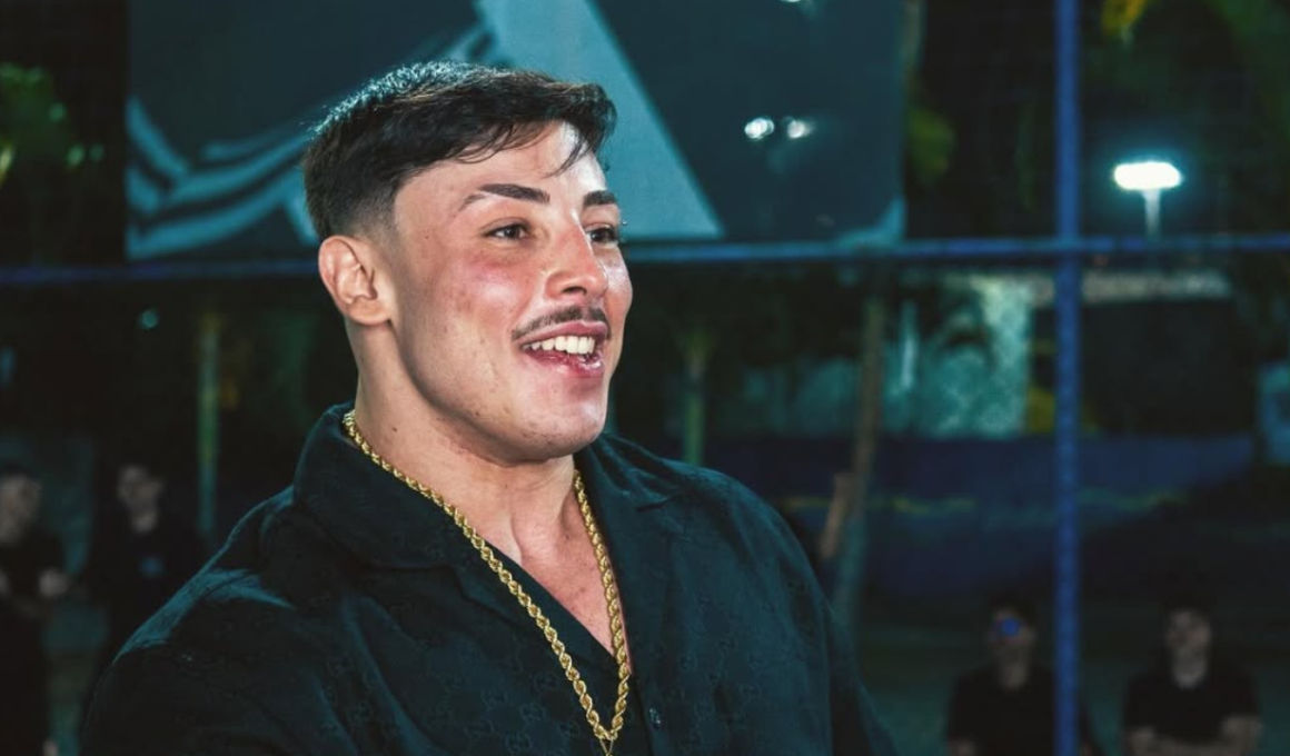 Gabriel Ganley sorrindo durante evento noturno em São Paulo. O influenciador e atleta aparece usando camisa preta, corrente dourada e cabelo curto estilizado, enquanto conversa em ambiente iluminado ao fundo.