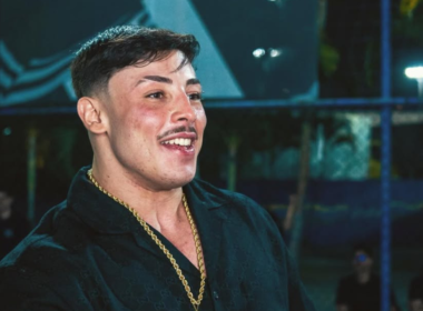 Gabriel Ganley sorrindo durante evento noturno em São Paulo. O influenciador e atleta aparece usando camisa preta, corrente dourada e cabelo curto estilizado, enquanto conversa em ambiente iluminado ao fundo.