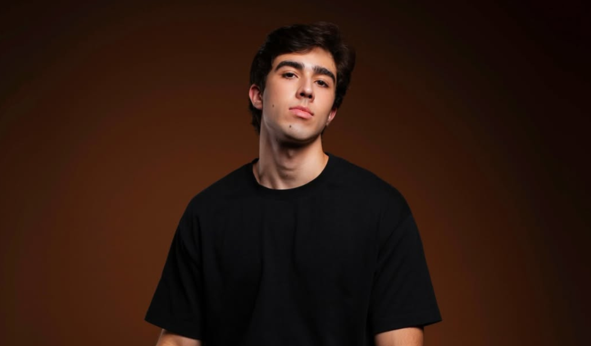 Homem jovem de cabelos escuros e camiseta preta posa diante de fundo marrom em ensaio fotográfico com iluminação dramática. Retrato de João Pedro Chaseliov, ator do musical de Paulo Gustavo, com expressão séria e visual minimalista.