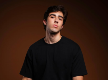 Homem jovem de cabelos escuros e camiseta preta posa diante de fundo marrom em ensaio fotográfico com iluminação dramática. Retrato de João Pedro Chaseliov, ator do musical de Paulo Gustavo, com expressão séria e visual minimalista.