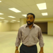 Homem de camisa cinza está em um amplo escritório vazio com iluminação fluorescente, remetendo ao cenário das Backrooms. A imagem apresenta um ambiente minimalista, com paredes bege, teto repleto de luzes e duas cadeiras ao fundo, transmitindo sensação de mistério, isolamento e suspense.