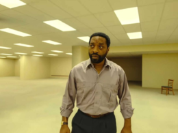 Homem de camisa cinza está em um amplo escritório vazio com iluminação fluorescente, remetendo ao cenário das Backrooms. A imagem apresenta um ambiente minimalista, com paredes bege, teto repleto de luzes e duas cadeiras ao fundo, transmitindo sensação de mistério, isolamento e suspense.