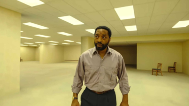 Homem de camisa cinza está em um amplo escritório vazio com iluminação fluorescente, remetendo ao cenário das Backrooms. A imagem apresenta um ambiente minimalista, com paredes bege, teto repleto de luzes e duas cadeiras ao fundo, transmitindo sensação de mistério, isolamento e suspense.