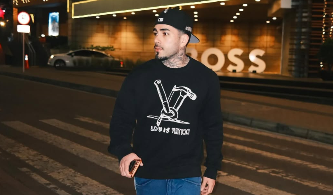 Streamer Brabox posa em uma rua iluminada durante a noite, usando boné preto, moletom estampado da Louis Vuitton e calça jeans azul. O criador de conteúdo aparece segurando um celular enquanto olha para o lado em frente a um prédio com iluminação moderna e letreiro ao fundo.