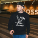 Streamer Brabox posa em uma rua iluminada durante a noite, usando boné preto, moletom estampado da Louis Vuitton e calça jeans azul. O criador de conteúdo aparece segurando um celular enquanto olha para o lado em frente a um prédio com iluminação moderna e letreiro ao fundo.