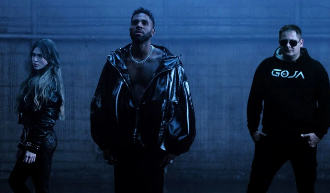 Foto promocional do clipe "Mi Chico" mostrando a cantora Melody de perfil, o cantor Jason Derulo ao centro sem camisa com jaqueta preta de látex, e o DJ Goja à direita vestindo moletom preto. O ambiente é escuro, com tons frios e névoa cenográfica.