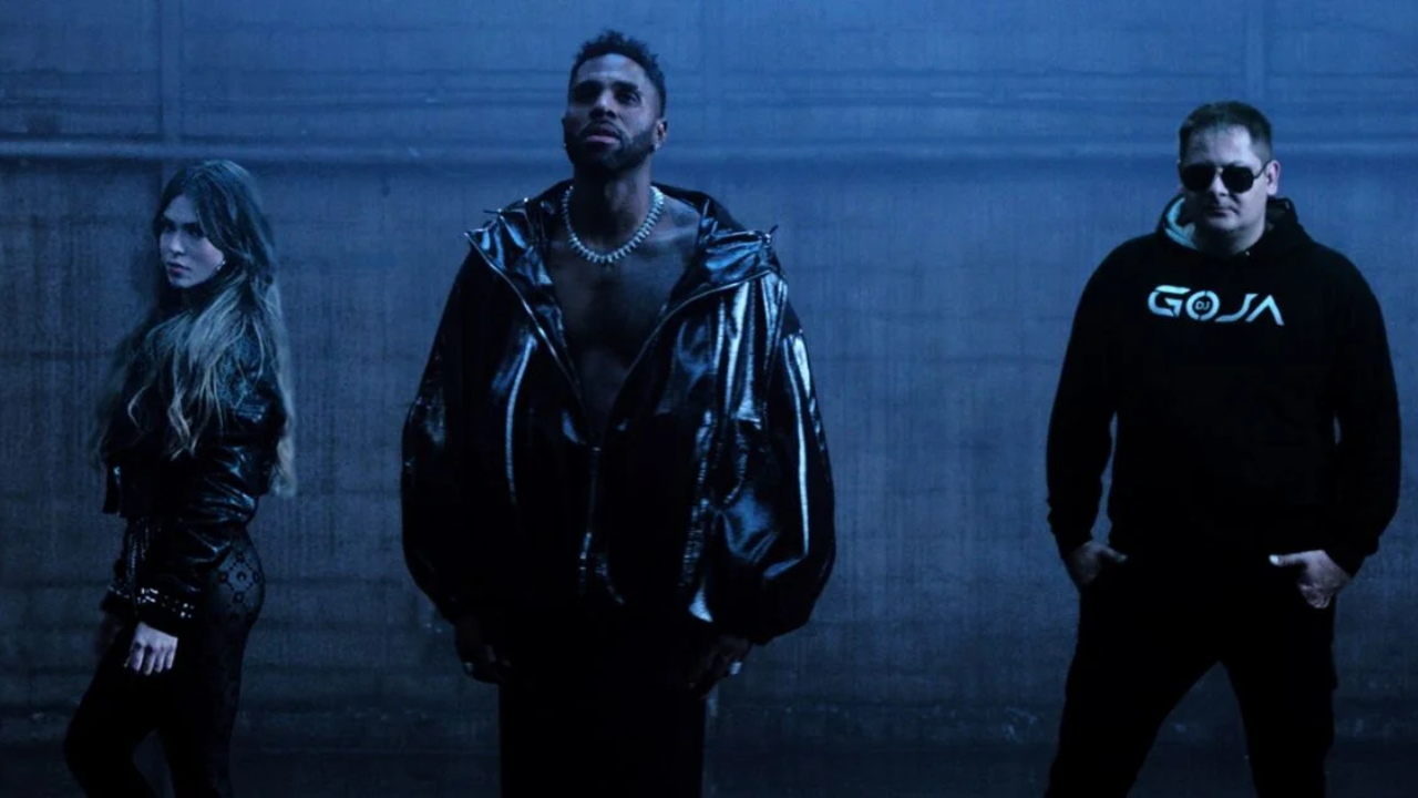 Foto promocional do clipe "Mi Chico" mostrando a cantora Melody de perfil, o cantor Jason Derulo ao centro sem camisa com jaqueta preta de látex, e o DJ Goja à direita vestindo moletom preto. O ambiente é escuro, com tons frios e névoa cenográfica.