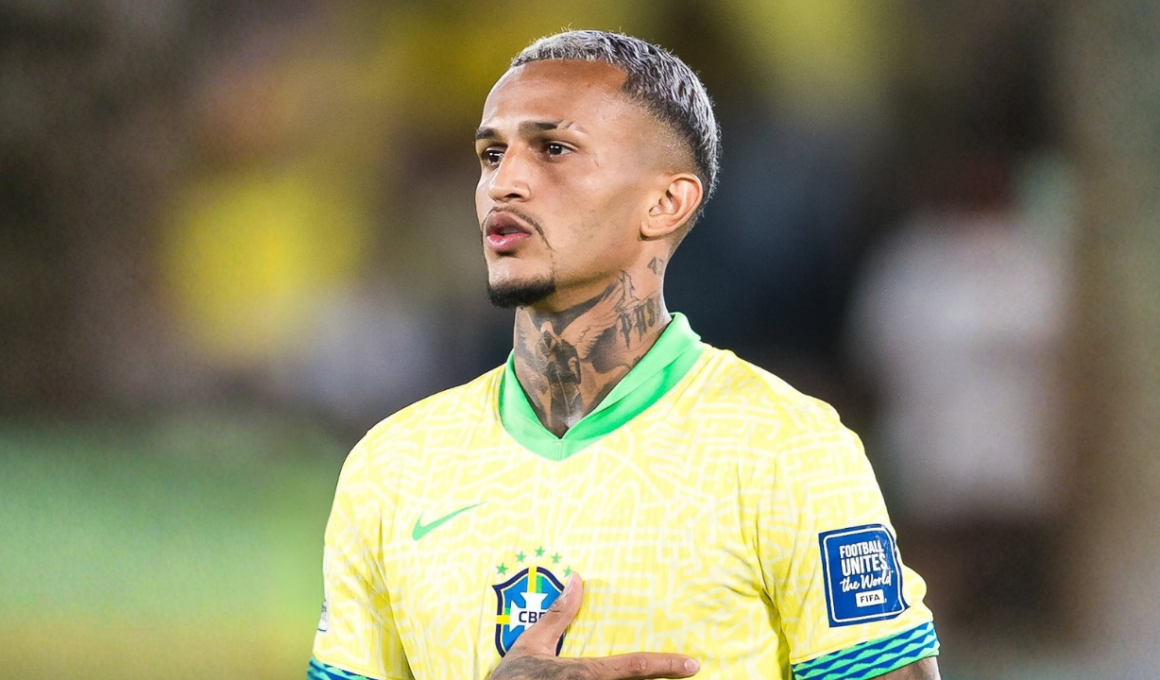 Wesley, jogador da Seleção Brasileira, aparece em close durante execução do hino nacional antes de partida oficial, usando camisa amarela da CBF, com cabelo platinado e tatuagens no pescoço em estádio ao fundo.