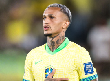 Wesley, jogador da Seleção Brasileira, aparece em close durante execução do hino nacional antes de partida oficial, usando camisa amarela da CBF, com cabelo platinado e tatuagens no pescoço em estádio ao fundo.