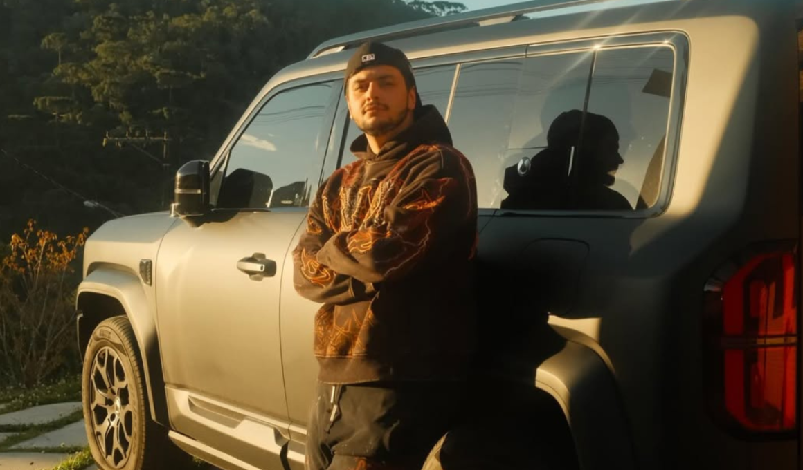 Streamer Gabepeixe posa encostado em um carro SUV branco durante o pôr do sol. O criador de conteúdo usa moletom escuro com detalhes em laranja e boné preto virado para trás, enquanto olha para a câmera com os braços cruzados. A imagem tem iluminação dourada e cenário externo com árvores ao fundo.