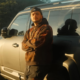 Streamer Gabepeixe posa encostado em um carro SUV branco durante o pôr do sol. O criador de conteúdo usa moletom escuro com detalhes em laranja e boné preto virado para trás, enquanto olha para a câmera com os braços cruzados. A imagem tem iluminação dourada e cenário externo com árvores ao fundo.