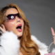 Alt text SEO: Wanessa Wolf posa em ensaio fotográfico com visual glamouroso, usando óculos escuros fashion, casaco branco de pelúcia e acessórios brilhantes. A influenciadora aparece com expressão surpresa, segurando uma bolsa rosa, em fundo cinza de estúdio, destacando seu estilo luxuoso e estética inspirada no universo pop e fashionista.