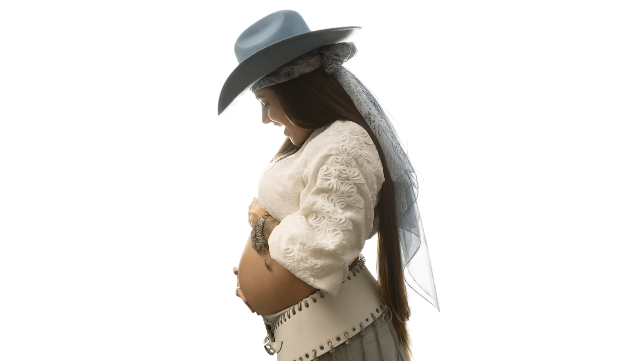 Lauana Prado posa de perfil exibindo a barriga de gravidez em ensaio fotográfico com fundo branco, usando chapéu azul-claro, blusa branca rendada e saia clara, em anúncio do nascimento do primeiro filho e lançamento de nova música.