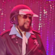 Victor Willis, vocalista do Village People como policial usando capacete, óculos escuros, gravata branca e jaqueta de couro preta durante apresentação em ambiente iluminado por luzes rosas e roxas, com cortinas de lantejoulas ao fundo.