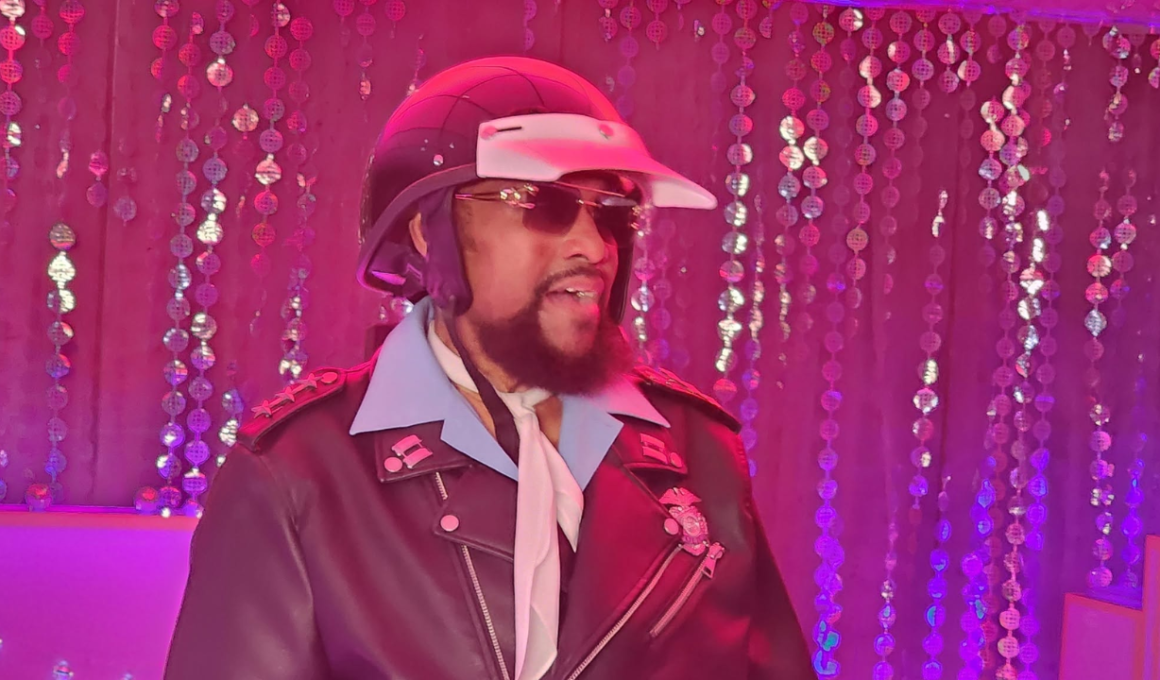 Victor Willis, vocalista do Village People como policial usando capacete, óculos escuros, gravata branca e jaqueta de couro preta durante apresentação em ambiente iluminado por luzes rosas e roxas, com cortinas de lantejoulas ao fundo.