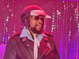 Victor Willis, vocalista do Village People como policial usando capacete, óculos escuros, gravata branca e jaqueta de couro preta durante apresentação em ambiente iluminado por luzes rosas e roxas, com cortinas de lantejoulas ao fundo.