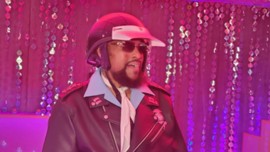 Victor Willis, vocalista do Village People como policial usando capacete, óculos escuros, gravata branca e jaqueta de couro preta durante apresentação em ambiente iluminado por luzes rosas e roxas, com cortinas de lantejoulas ao fundo.