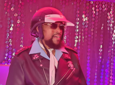 Victor Willis, vocalista do Village People como policial usando capacete, óculos escuros, gravata branca e jaqueta de couro preta durante apresentação em ambiente iluminado por luzes rosas e roxas, com cortinas de lantejoulas ao fundo.