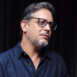 Lúcio Mauro Filho usa óculos e camisa azul durante entrevista sobre diagnóstico de câncer no intestino.