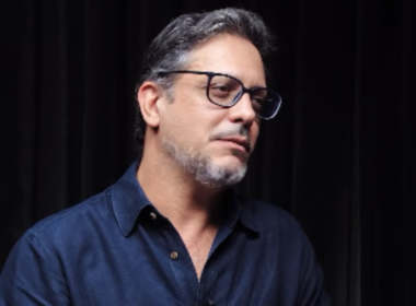 Lúcio Mauro Filho usa óculos e camisa azul durante entrevista sobre diagnóstico de câncer no intestino.