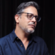Lúcio Mauro Filho usa óculos e camisa azul durante entrevista sobre diagnóstico de câncer no intestino.