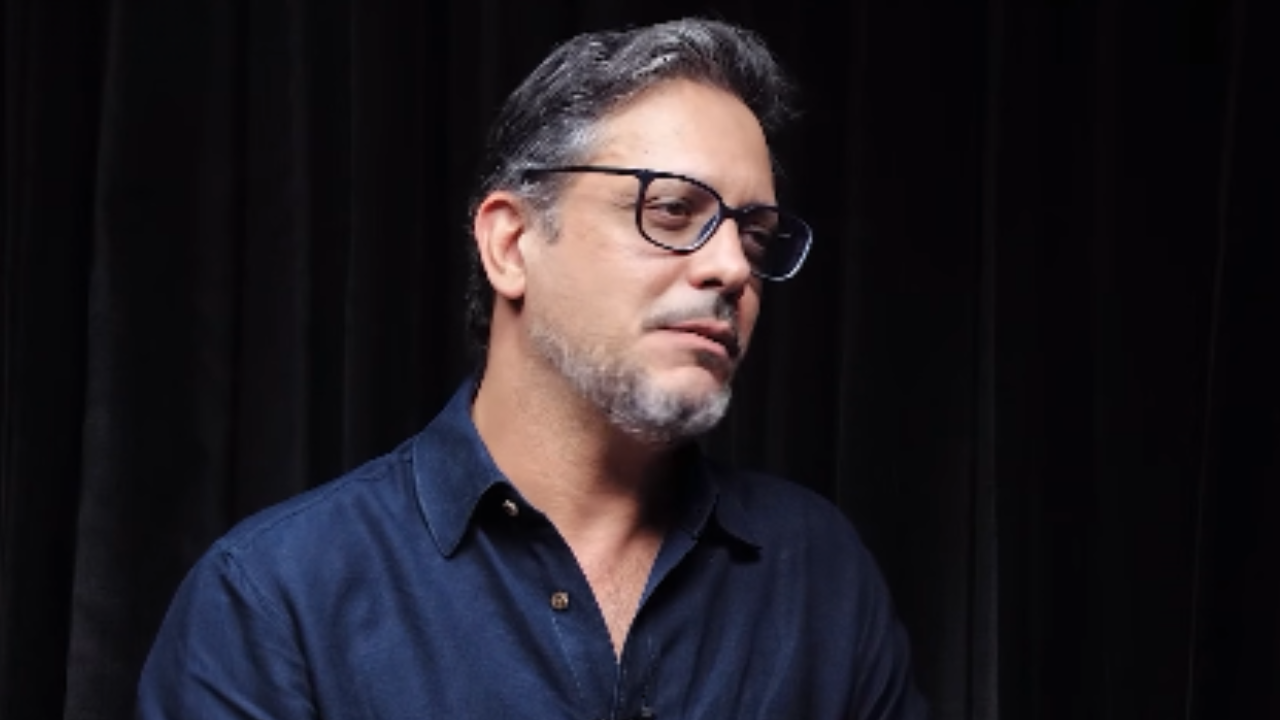 Lúcio Mauro Filho usa óculos e camisa azul durante entrevista sobre diagnóstico de câncer no intestino.