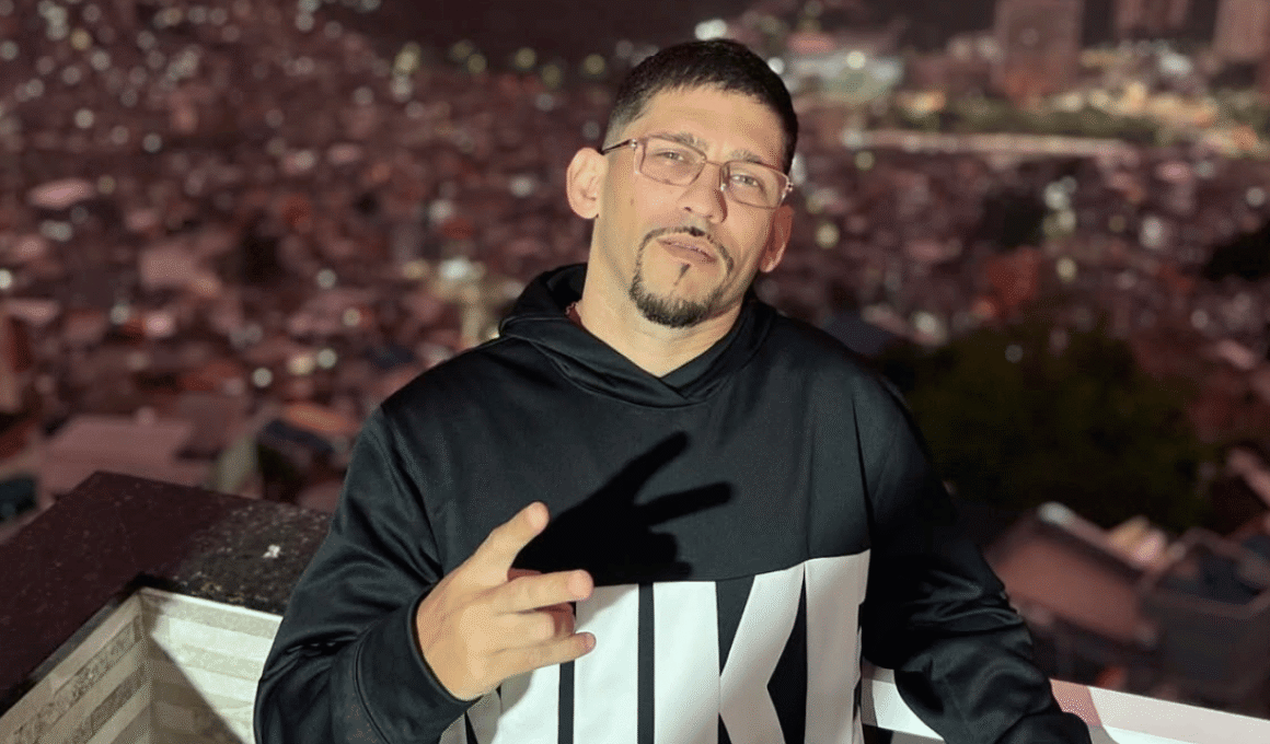 MC Orelha, um homem de cabelo curto, óculos e cavanhaque usando moletom preto com estampa, fazendo sinal de paz com a mão em varanda com vista noturna para cidade ilumina ao fundo.
