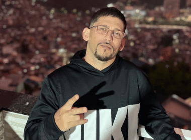 MC Orelha, um homem de cabelo curto, óculos e cavanhaque usando moletom preto com estampa, fazendo sinal de paz com a mão em varanda com vista noturna para cidade ilumina ao fundo.
