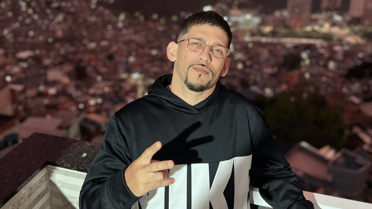 MC Orelha, um homem de cabelo curto, óculos e cavanhaque usando moletom preto com estampa, fazendo sinal de paz com a mão em varanda com vista noturna para cidade ilumina ao fundo.