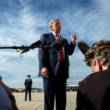 Donald Trump de terno azul-marinho e gravata vermelha fala com jornalistas durante uma entrevista ao ar livre, enquanto microfones e câmeras registram o momento. Ao fundo, há uma pista de aeroporto e um avião estacionado sob um céu azul.