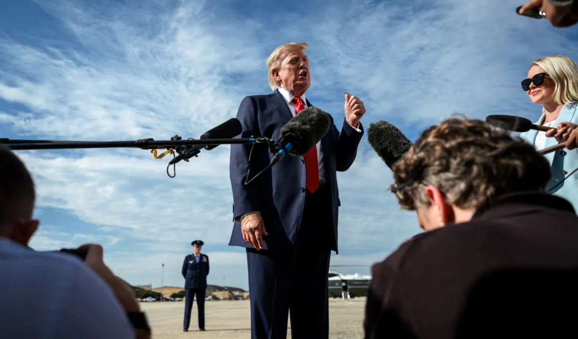 Donald Trump de terno azul-marinho e gravata vermelha fala com jornalistas durante uma entrevista ao ar livre, enquanto microfones e câmeras registram o momento. Ao fundo, há uma pista de aeroporto e um avião estacionado sob um céu azul.