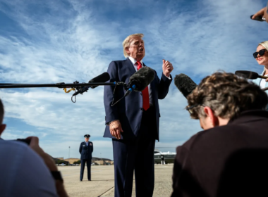 Donald Trump de terno azul-marinho e gravata vermelha fala com jornalistas durante uma entrevista ao ar livre, enquanto microfones e câmeras registram o momento. Ao fundo, há uma pista de aeroporto e um avião estacionado sob um céu azul.